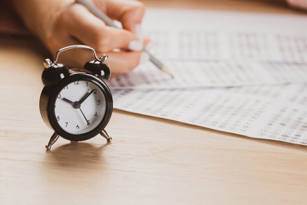 Black alarm clock on wooden desk beside person writing notes, representing study time for FRACP exam preparation.